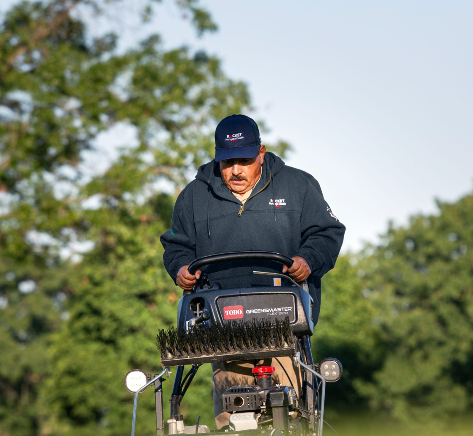 Groundskeeper operating equipment in an embroidered navy hoodie and cap